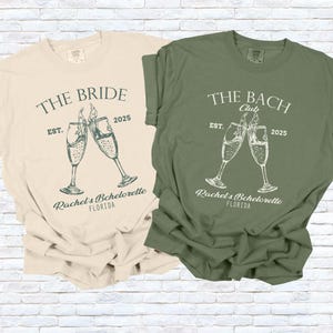 Pu&ograve; includere: Due magliette, una color crema e una verde oliva, con grafiche abbinate. La maglietta color crema dice "The Bride" con calici di champagne e "Rachel's Bachelorette, Florida". La maglietta verde dice "The Bach Club" con calici di champagne e "Rachel's Bachelorette, Florida".