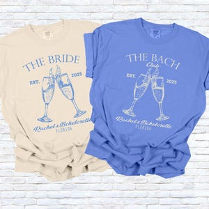 Pu&ograve; includere: Due magliette a maniche corte, una color crema e una blu, con grafiche abbinate. La maglietta color crema dice "THE BRIDE" con calici di champagne e "Rachel's Bachelorette FLORIDA" e "EST. 2025". La maglietta blu dice "THE BACH Club".