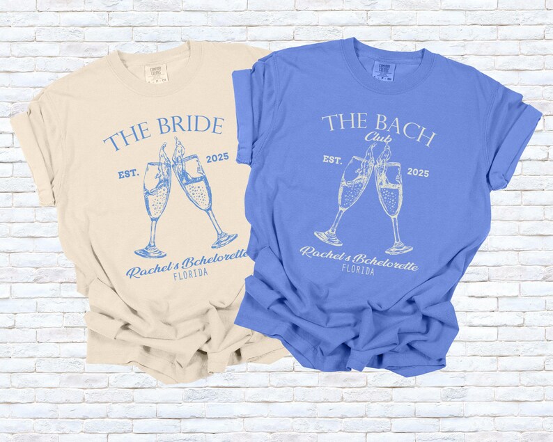 Pu&ograve; includere: Due magliette a maniche corte, una color crema e una blu, con grafiche abbinate. La maglietta color crema dice "THE BRIDE" con calici di champagne e "Rachel's Bachelorette FLORIDA" e "EST. 2025". La maglietta blu dice "THE BACH Club".