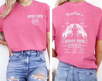 May include: A pink t-shirt with the text "BRIDE CREW" on the front and a rodeo-themed design on the back. The back of the shirt features the text "Everlyn's Rodeo Tour Austin, TX" with a graphic of two riders on horses.