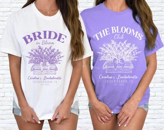 May include: A white t-shirt with purple text reading BRIDE in Bloom and a purple floral graphic, and a light purple t-shirt with white text reading THE BLOOMS Club and a white floral graphic. Both shirts feature Caroline's Bachelorette, FREDERICKSBURG, TX, EST 2025.
