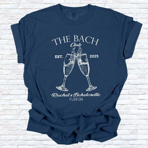 Pu&ograve; includere: T-shirt blu navy con testo e grafica bianchi. Il testo recita "THE BACH Club EST. 2025" sopra due calici di champagne che si toccano. Sotto i calici c'&egrave; "Rachel's Bachelorette FLORIDA".