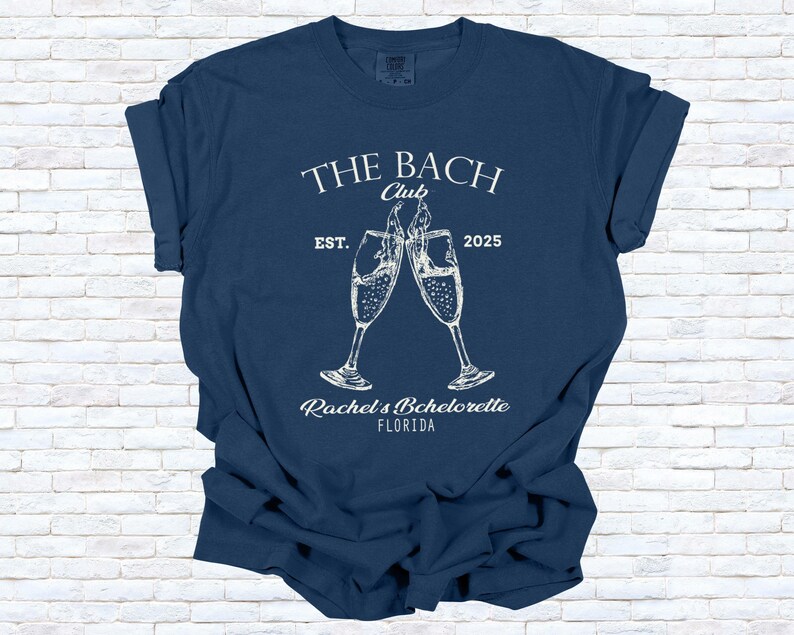 Pu&ograve; includere: T-shirt blu navy con testo e grafica bianchi. Il testo recita "THE BACH Club EST. 2025" sopra due calici di champagne che si toccano. Sotto i calici c'&egrave; "Rachel's Bachelorette FLORIDA".