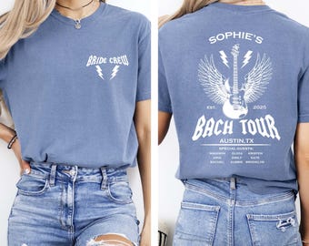 May include: A blue t-shirt with white text that reads "BRIDE CREW" and two lightning bolts on the front. The back of the shirt features a guitar with wings and the text "SOPHIE'S BACH TOUR AUSTIN, TX 2025".
