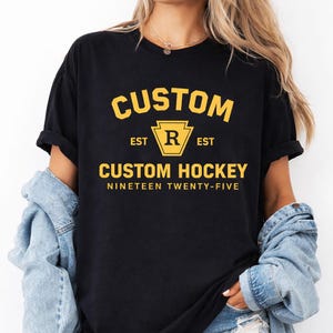May include: Black t-shirt with a gold graphic that reads "CUSTOM" above "CUSTOM HOCKEY" and "NINETEEN TWENTY-FIVE." The graphic includes a stylized "R" inside a gold keystone shape. The shirt is worn by a person.
