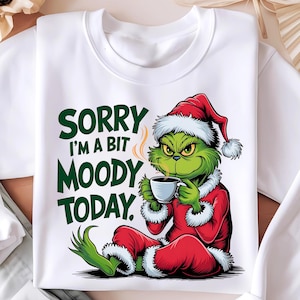 May include: White sweatshirt with a green Grinch character wearing a red Santa hat and holding a cup of coffee. The text "Sorry I'm a bit Moody Today." is printed in green on the shirt.