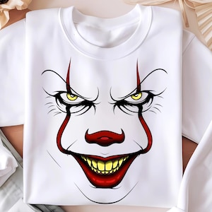 May include: White sweatshirt featuring a graphic of a scary clown face. The clown has yellow eyes, a red nose, and a wide red mouth with yellow teeth. Red lines extend from the eyes and mouth.
