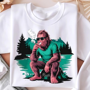 Peut inclure: Un t-shirt blanc avec un motif représentant un Bigfoot de dessin animé portant des lunettes de soleil et fumant une cigarette. Le Bigfoot est assis sur un rocher dans un décor de forêt avec un lac en arrière-plan.