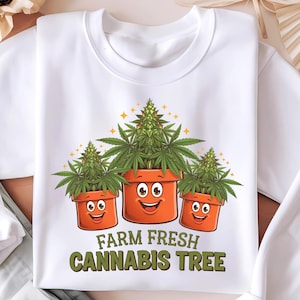 以下が含まれることがあります： 笑顔の顔をした鉢植えのマリファナ植物3つが描かれた白いスウェットシャツ。植物の下には「Farm Fresh Cannabis Tree」という文字が印刷されています。