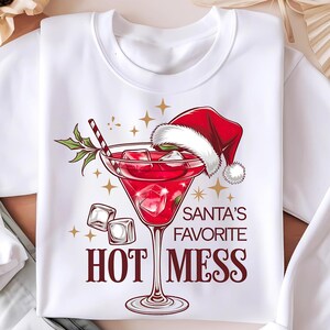 Puede incluir: Sudadera blanca de Navidad con una copa de martini roja con hielo y un gorro de Papá Noel rojo. El texto dice "El favorito de Papá Noel: Un desastre caliente".