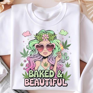 Peut inclure: Sweat-shirt blanc avec une illustration de dessin animé d'une femme aux cheveux verts et roses portant des lunettes de soleil en forme de cœur et une couronne de fleurs. Elle tient un joint allumé et il y a des feuilles de marijuana vertes autour d'elle. Le texte "Baked & Beautiful" est imprimé en lettres roses et vertes sous l'illustration.