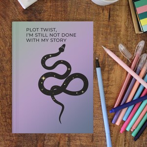 May include: A purple and blue gradient notebook with the text "PLOT TWIST, I'M STILL NOT DONE WITH MY STORY" and a black snake with gold stars on it.