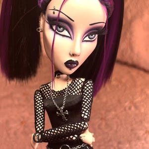 May include: A goth-inspired doll with purple and black hair, wearing a black mesh top, a black skirt, and a silver chain necklace with a cross pendant. The doll has dark makeup, including black eyeliner, purple eyeshadow, and dark lipstick.