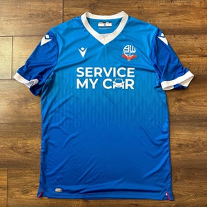 May include: A blue soccer jersey with white accents and the team logo. The jersey has the words "SERVICE MY CAR" printed on the front.