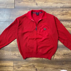 May include: A bright red long-sleeved sweater with a quarter-zip collar. The sweater has a ribbed texture and a small logo patch on the chest. The collar is a darker shade of red. The sweater is laid flat on a wooden surface.