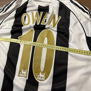 K&ouml;nnte beinhalten: Ein schwarz-wei&szlig; gestreiftes Fu&szlig;balltrikot mit dem Namen "OWEN" und der Nummer "10" in Gold. Ein Ma&szlig;band liegt &uuml;ber dem Trikot und zeigt die L&auml;nge in Zentimetern an.
