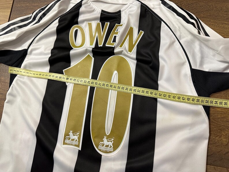 K&ouml;nnte beinhalten: Ein schwarz-wei&szlig; gestreiftes Fu&szlig;balltrikot mit dem Namen "OWEN" und der Nummer "10" in Gold. Ein Ma&szlig;band liegt &uuml;ber dem Trikot und zeigt die L&auml;nge in Zentimetern an.