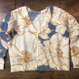 May include: Cream-colored cardigan sweater with a V-neck and button closure. The sweater features a floral pattern in shades of white and gold against a blue background. The label is visible at the neckline.