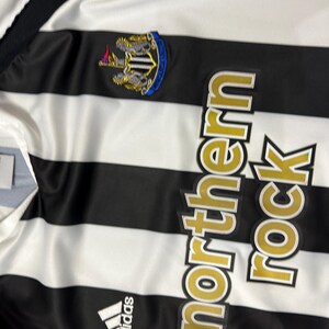 K&ouml;nnte beinhalten: Ein schwarz-wei&szlig; gestreiftes Fu&szlig;balltrikot mit dem Text "northern rock" in goldfarbenen Buchstaben. Das Trikot zeigt das Adidas-Logo und ein Teamwappen mit blauem Rand.