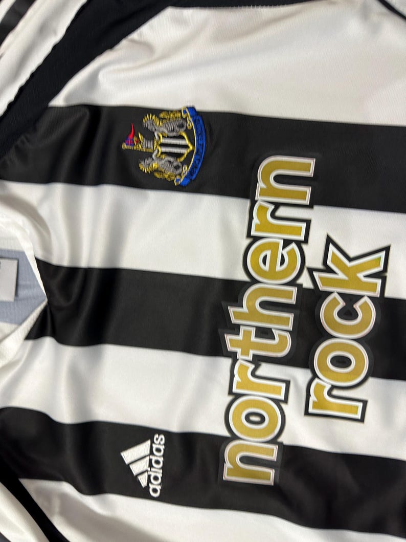 K&ouml;nnte beinhalten: Ein schwarz-wei&szlig; gestreiftes Fu&szlig;balltrikot mit dem Text "northern rock" in goldfarbenen Buchstaben. Das Trikot zeigt das Adidas-Logo und ein Teamwappen mit blauem Rand.
