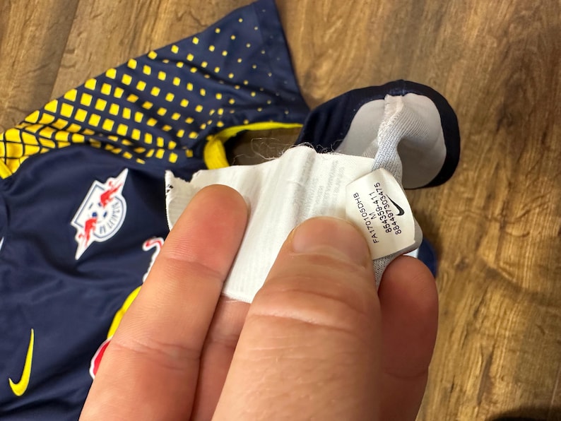 May include: A navy blue soccer jersey with yellow accents and a red bull logo. The jersey has a white tag with the text "FA1T01030CH1B M 854359-411 884467303475".