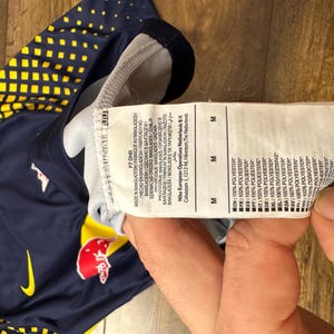 May include: A navy blue and yellow soccer jersey with a red bull logo and a white Nike swoosh. The jersey has a size tag that reads "M" and lists the fabric composition as 100% polyester.