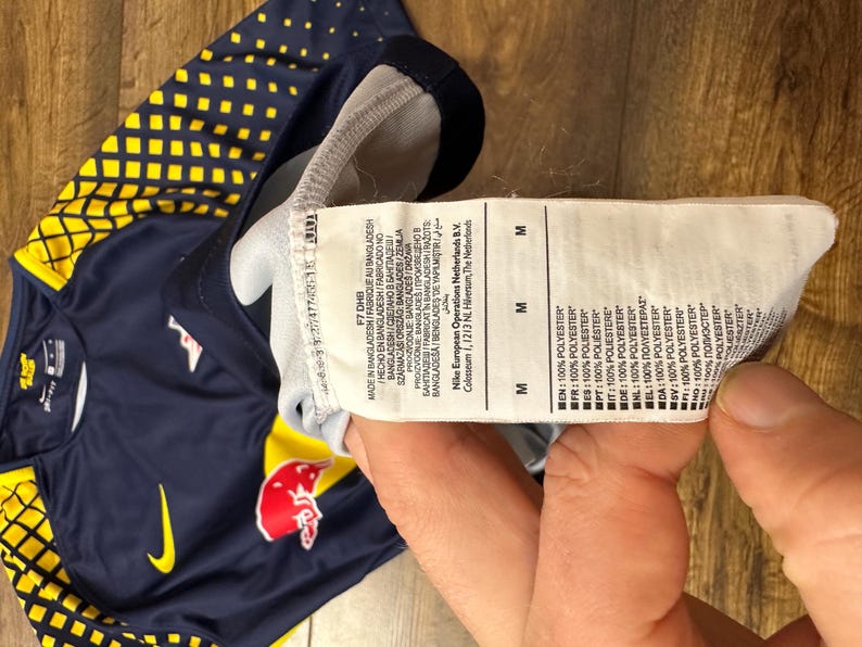 May include: A navy blue and yellow soccer jersey with a red bull logo and a white Nike swoosh. The jersey has a size tag that reads "M" and lists the fabric composition as 100% polyester.