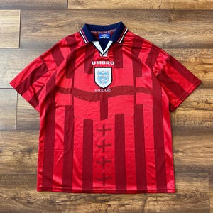 以下が含まれることがあります： アンブロのロゴとイングランドの紋章が付いた赤と白のストライプのサッカーユニフォーム。ユニフォームは白の縁取りのある青い襟です。