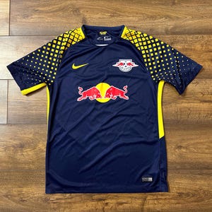 May include: A navy blue soccer jersey with yellow accents and the Red Bull logo. The jersey has a Nike swoosh logo on the left chest and the Red Bull logo on the right chest.