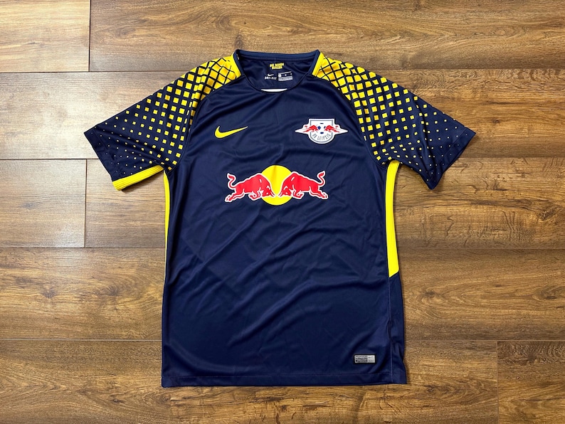 May include: A navy blue soccer jersey with yellow accents and the Red Bull logo. The jersey has a Nike swoosh logo on the left chest and the Red Bull logo on the right chest.
