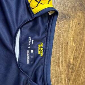 May include: A navy blue soccer jersey with yellow and black geometric pattern. The jersey has a white Nike swoosh logo and the text "DRI-FIT" and "DIE ROTEN BULLEN" on the tag. The tag also indicates the size is "M".