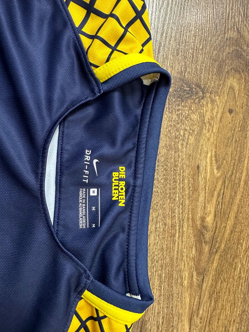 May include: A navy blue soccer jersey with yellow and black geometric pattern. The jersey has a white Nike swoosh logo and the text "DRI-FIT" and "DIE ROTEN BULLEN" on the tag. The tag also indicates the size is "M".