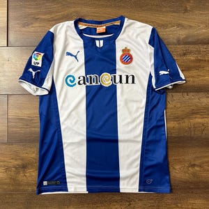 Puede incluir: Una camiseta de fútbol a rayas azules y blancas con el logotipo del equipo y la palabra "camcun" en la parte delantera.