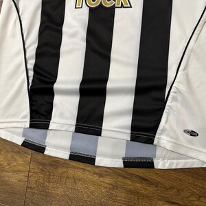 K&ouml;nnte beinhalten: Wei&szlig;-schwarz gestreiftes Sporttrikot. Das Trikot hat einen Rundhalsausschnitt und kurze &Auml;rmel. Das Wort "ROCK" ist oben sichtbar. Das Trikot hat ein "ClimaCool"-Logo.