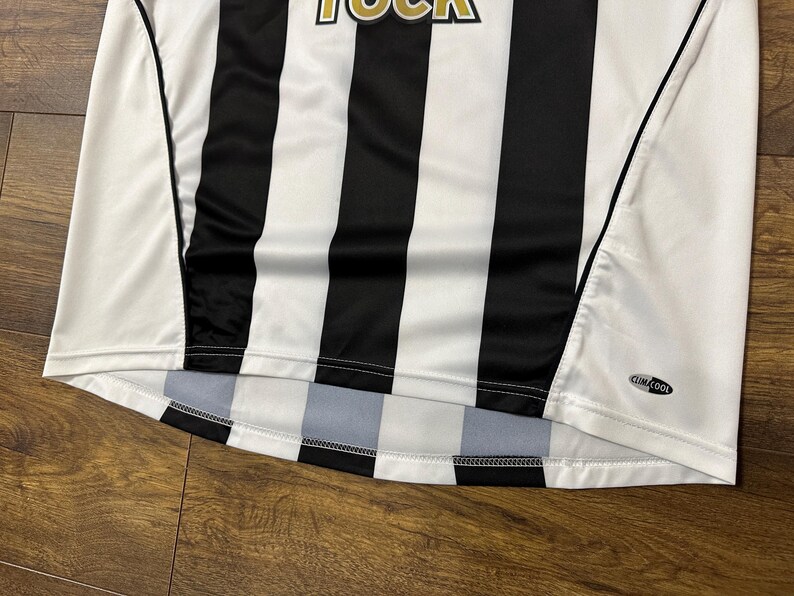 K&ouml;nnte beinhalten: Wei&szlig;-schwarz gestreiftes Sporttrikot. Das Trikot hat einen Rundhalsausschnitt und kurze &Auml;rmel. Das Wort "ROCK" ist oben sichtbar. Das Trikot hat ein "ClimaCool"-Logo.
