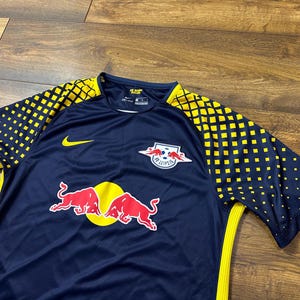 May include: A navy blue soccer jersey with yellow and black accents. The jersey has the Red Bull logo and the text "RB Leipzig" on the front. The jersey also has a Nike logo on the left sleeve.