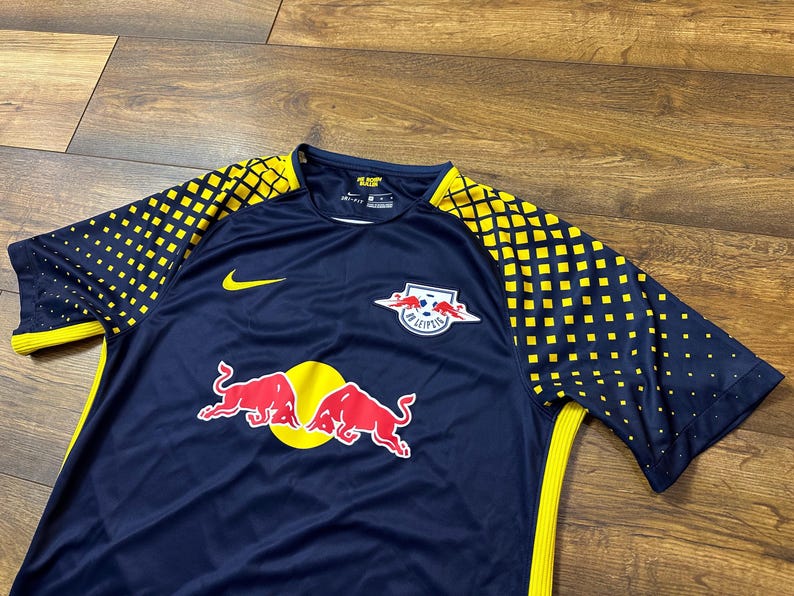 May include: A navy blue soccer jersey with yellow and black accents. The jersey has the Red Bull logo and the text "RB Leipzig" on the front. The jersey also has a Nike logo on the left sleeve.