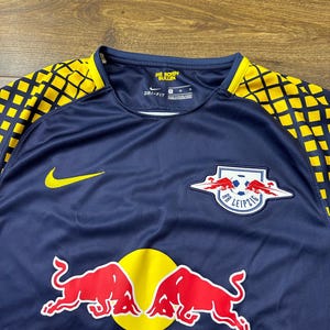 May include: A navy blue soccer jersey with yellow and black accents. The jersey features the team logo, "RB Leipzig", with two red bulls and a soccer ball. The Nike logo is on the left side of the chest. The text "Die Roten Bullen" is on the back of the neck.
