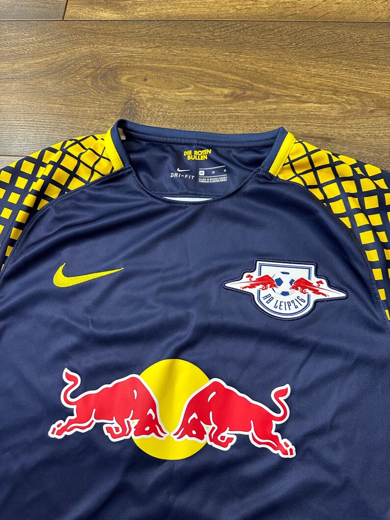 May include: A navy blue soccer jersey with yellow and black accents. The jersey features the team logo, "RB Leipzig", with two red bulls and a soccer ball. The Nike logo is on the left side of the chest. The text "Die Roten Bullen" is on the back of the neck.