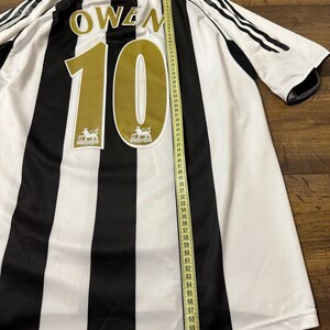 K&ouml;nnte beinhalten: Ein schwarz-wei&szlig; gestreiftes Fu&szlig;balltrikot mit dem Namen "OWEN" und der Nummer "10" in Gold. Das Trikot hat ein Ma&szlig;band daneben, das seine L&auml;nge anzeigt. Das Trikot hat das Premier League-Logo.