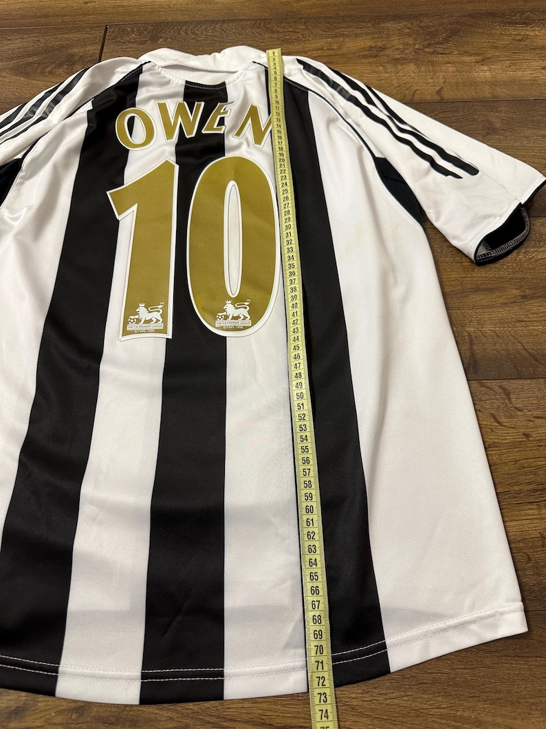 K&ouml;nnte beinhalten: Ein schwarz-wei&szlig; gestreiftes Fu&szlig;balltrikot mit dem Namen "OWEN" und der Nummer "10" in Gold. Das Trikot hat ein Ma&szlig;band daneben, das seine L&auml;nge anzeigt. Das Trikot hat das Premier League-Logo.