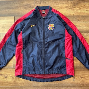 Puede incluir: Una chaqueta cortavientos azul marino y roja con cierre de cremallera. La chaqueta tiene un logotipo de Nike en el pecho izquierdo y un escudo del FC Barcelona en el pecho derecho.