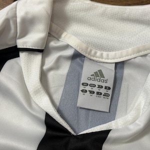 K&ouml;nnte beinhalten: Nahaufnahme eines wei&szlig;-schwarz gestreiften Adidas-Fu&szlig;balltrikots. Das Trikot hat einen V-Ausschnitt und das Adidas-Logo ist auf einem Etikett zu sehen. Das Trikot ist in der Gr&ouml;&szlig;e M.