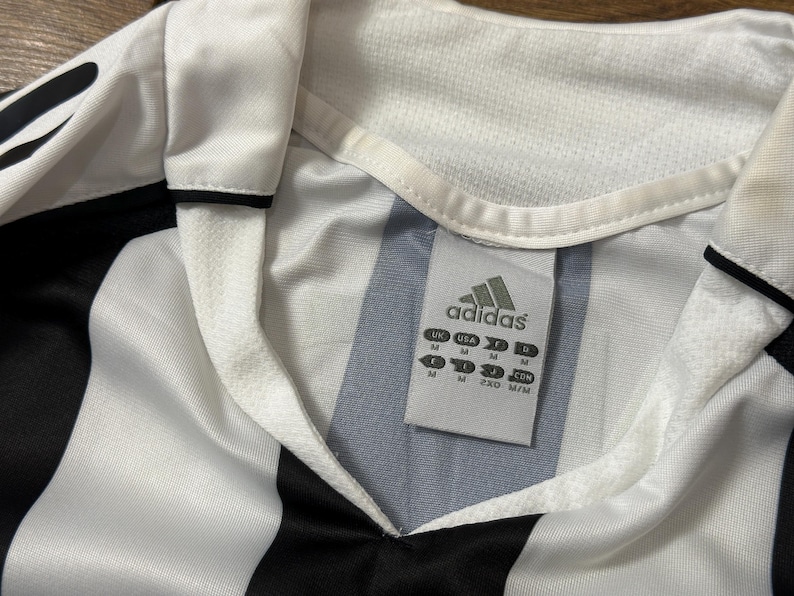 K&ouml;nnte beinhalten: Nahaufnahme eines wei&szlig;-schwarz gestreiften Adidas-Fu&szlig;balltrikots. Das Trikot hat einen V-Ausschnitt und das Adidas-Logo ist auf einem Etikett zu sehen. Das Trikot ist in der Gr&ouml;&szlig;e M.