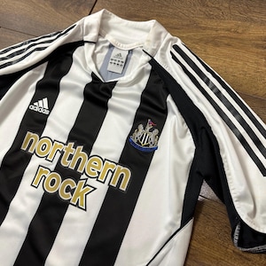 K&ouml;nnte beinhalten: Schwarz-wei&szlig; gestreiftes Fu&szlig;balltrikot mit goldenem "northern rock"-Schriftzug. Das Trikot hat das Adidas-Logo und das Vereinswappen. Die &Auml;rmel haben schwarze Akzente und wei&szlig;e Streifen.