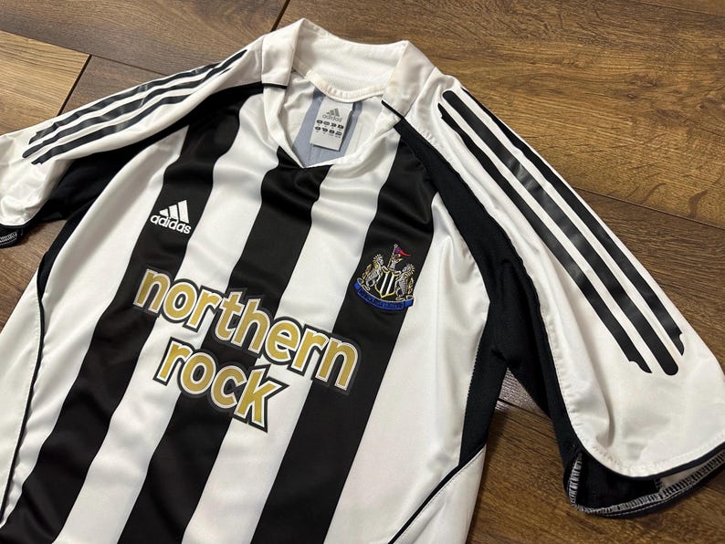K&ouml;nnte beinhalten: Schwarz-wei&szlig; gestreiftes Fu&szlig;balltrikot mit goldenem "northern rock"-Schriftzug. Das Trikot hat das Adidas-Logo und das Vereinswappen. Die &Auml;rmel haben schwarze Akzente und wei&szlig;e Streifen.
