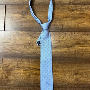 May include: A light blue tie with a white and light blue patterned design. The tie has a small blue tag attached to it.
