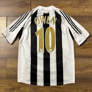 K&ouml;nnte beinhalten: Schwarz-wei&szlig; gestreiftes Fu&szlig;balltrikot mit dem Namen "OWEN" und der Nummer "10" in Gold. Das Trikot hat schwarze Streifen auf den &Auml;rmeln und das Premier League-Logo.