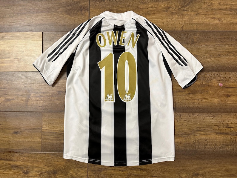 K&ouml;nnte beinhalten: Schwarz-wei&szlig; gestreiftes Fu&szlig;balltrikot mit dem Namen "OWEN" und der Nummer "10" in Gold. Das Trikot hat schwarze Streifen auf den &Auml;rmeln und das Premier League-Logo.