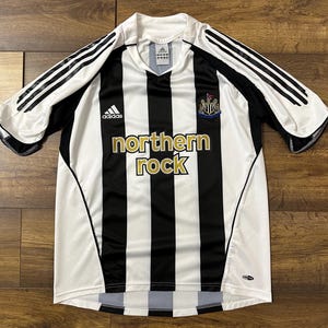 K&ouml;nnte beinhalten: Ein schwarz-wei&szlig; gestreiftes Fu&szlig;balltrikot mit dem Schriftzug "northern rock" in Gold. Das Trikot hat das Adidas-Logo und das Wappen von Newcastle United. Die &Auml;rmel sind wei&szlig; mit schwarzen Streifen.